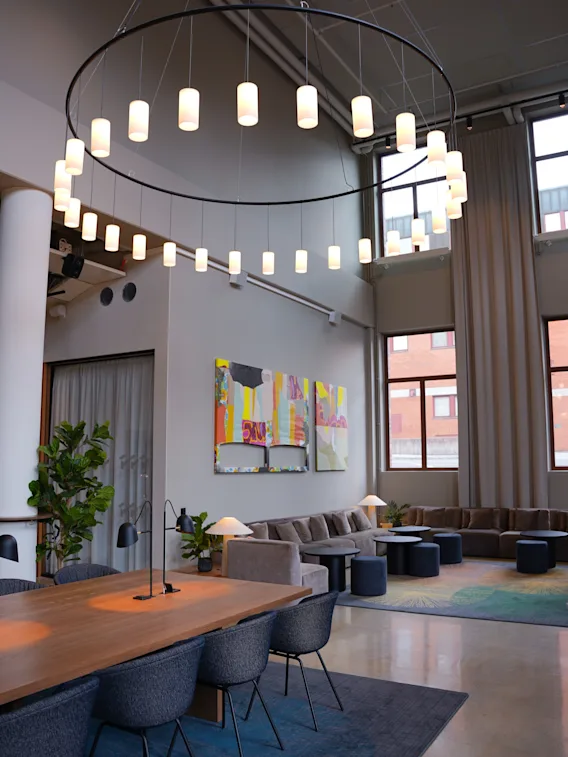 En hemtrevlig hotellounge med en stor cirkulär ljuskrona, modern konst på väggarna, mjuka soffor och ett långt träbord med blå stolar. Höga fönster släpper in naturligt ljus och skapar en inbjudande atmosfär.