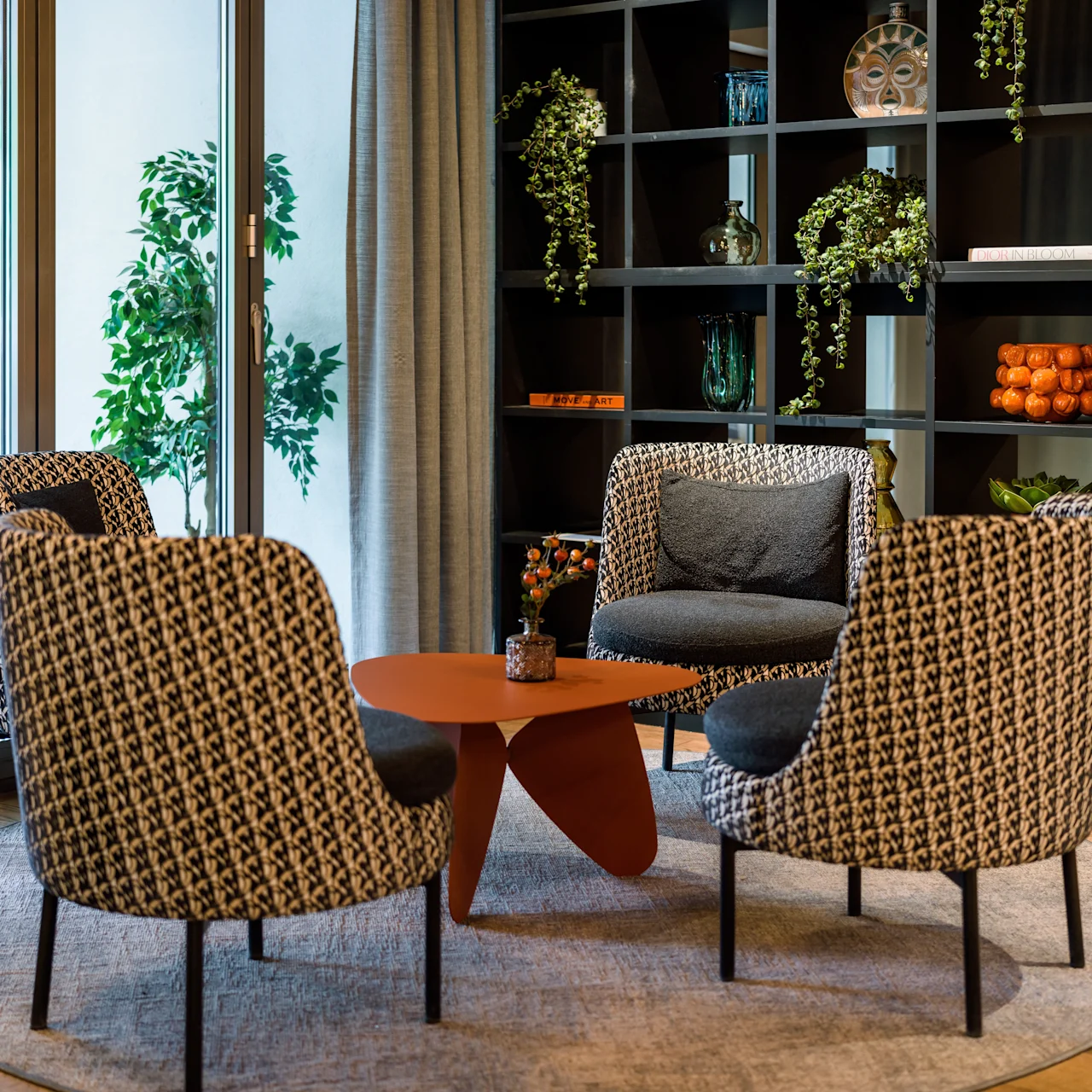 Four patterned armchairs encircle a modern, orange coffee table on a round rug. Shelving with plants and decor adorns the wall, near large glass windows with curtains.