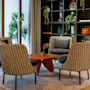 Four patterned armchairs encircle a modern, orange coffee table on a round rug. Shelving with plants and decor adorns the wall, near large glass windows with curtains.