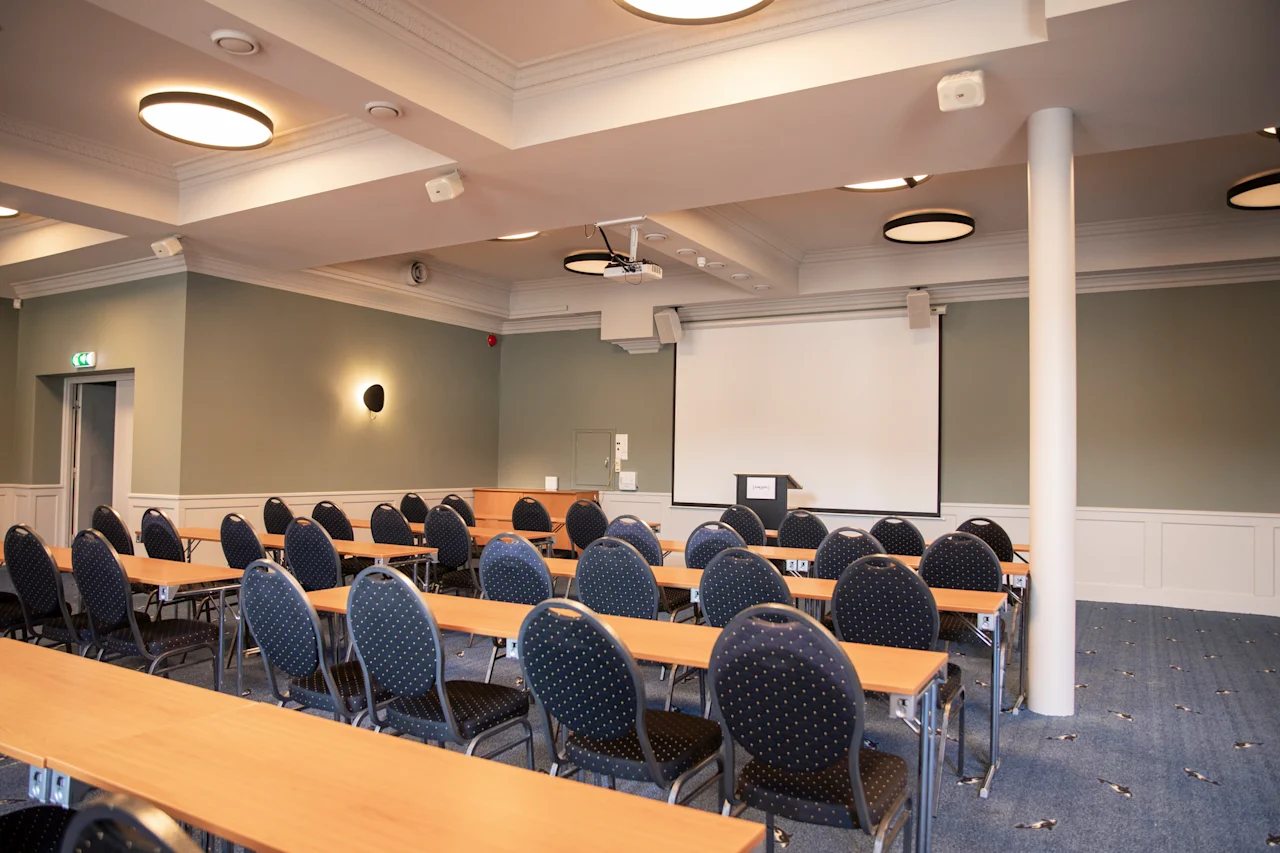 Et konferencelokale med rækker af borde og stole vendt mod en projektorskærm og et podium, klar til en præsentation.