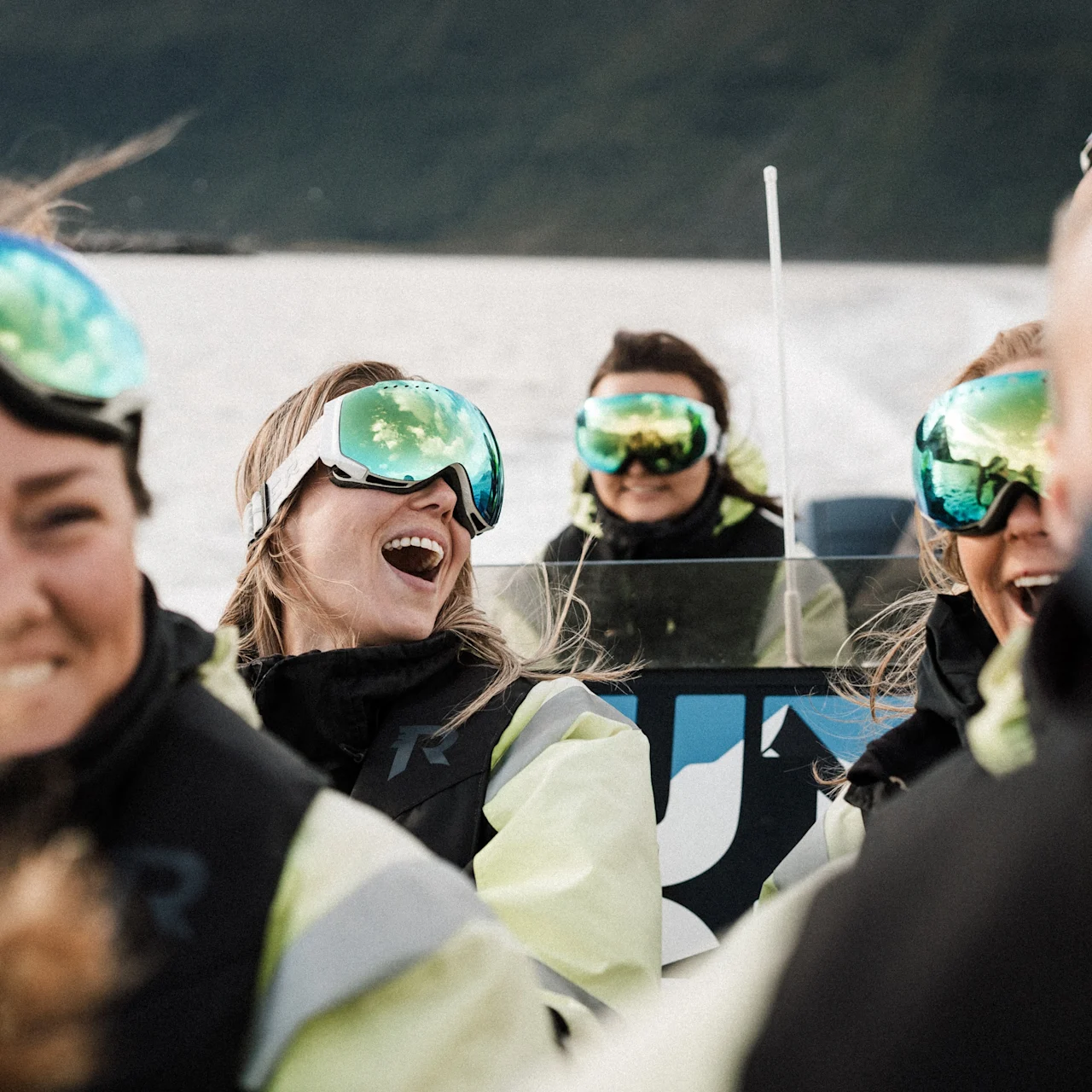 Människor i skidglasögon skrattar och njuter av en utomhusaktivitet på vattnet, med himlens reflektioner i glasögonen.