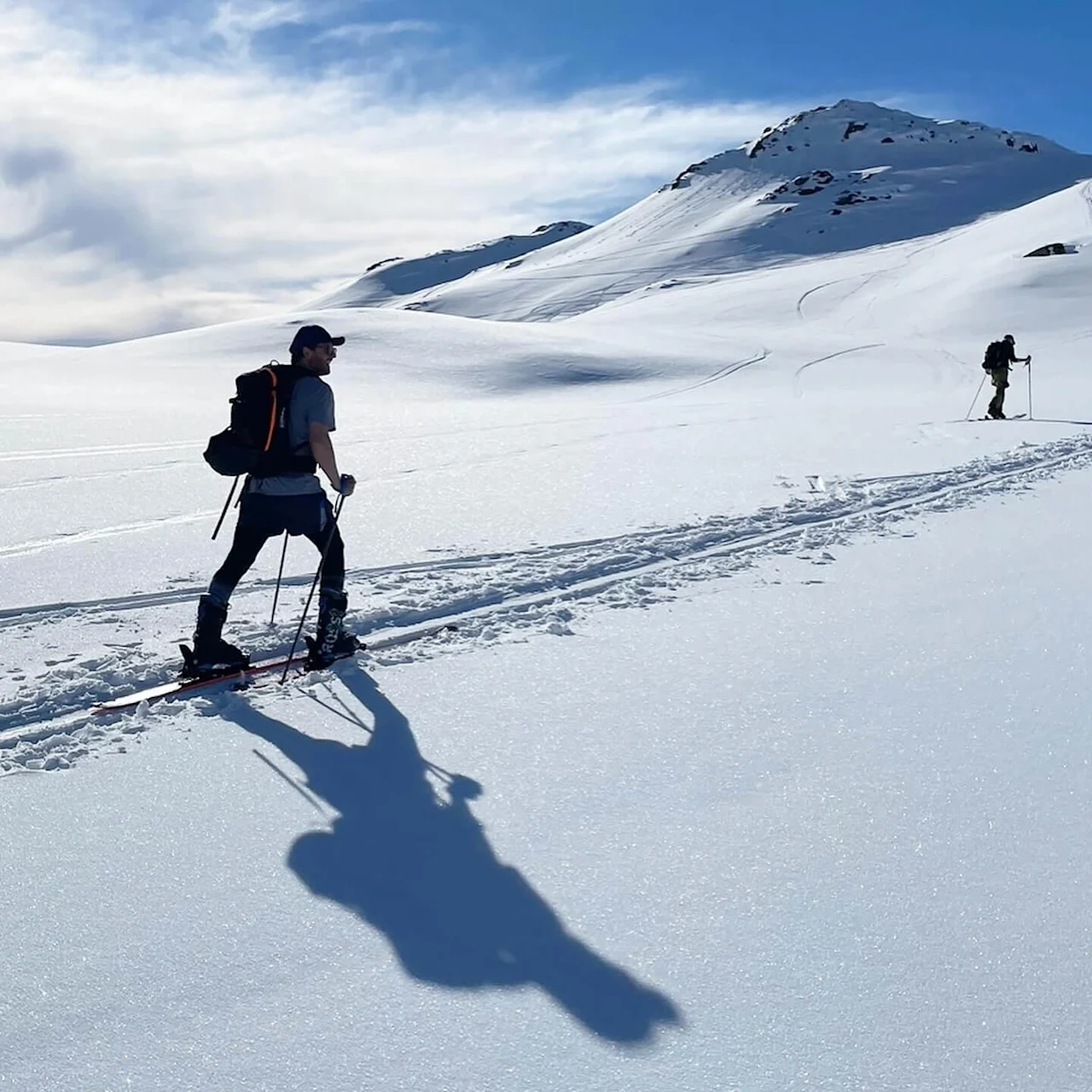 A person skiing uphill in snowy mountains under a clear blue sky. Another skier is visible in the distance, with shadows cast on the snow.