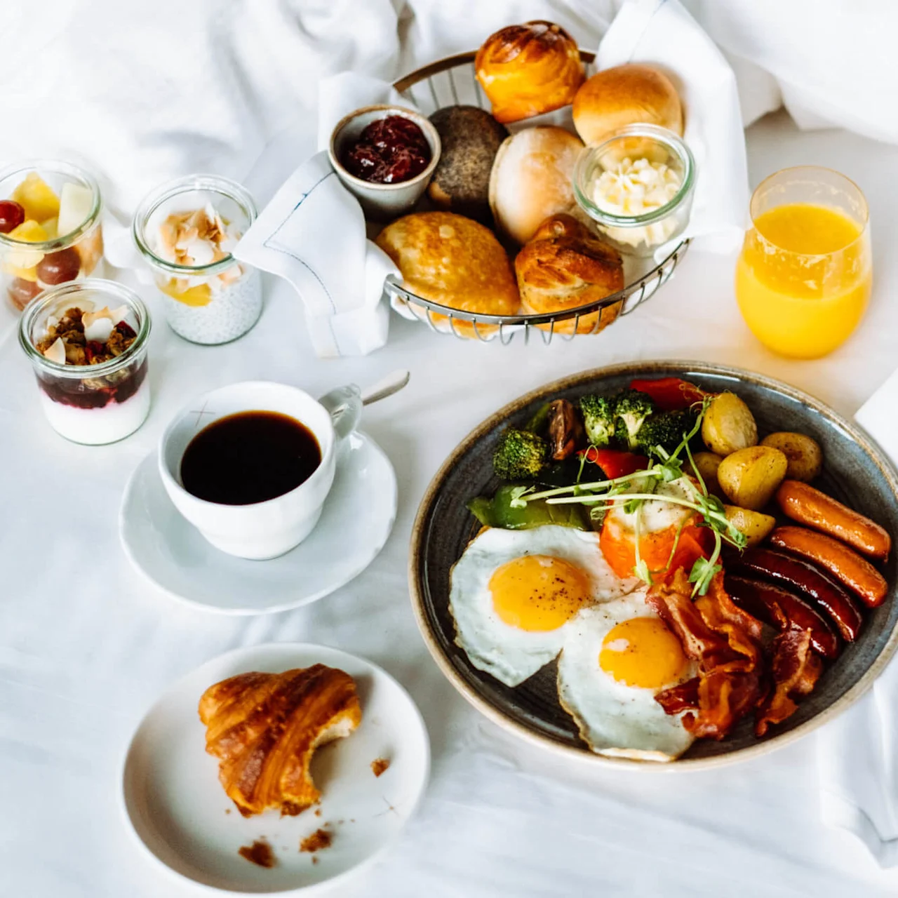 Frokost lagt opp på en seng, med egg, bacon, pølser, grønnsaker og poteter på en tallerken; bakverk i en kurv, med kaffe, appelsinjuice, yoghurt, frukt og en croissant ved siden av.