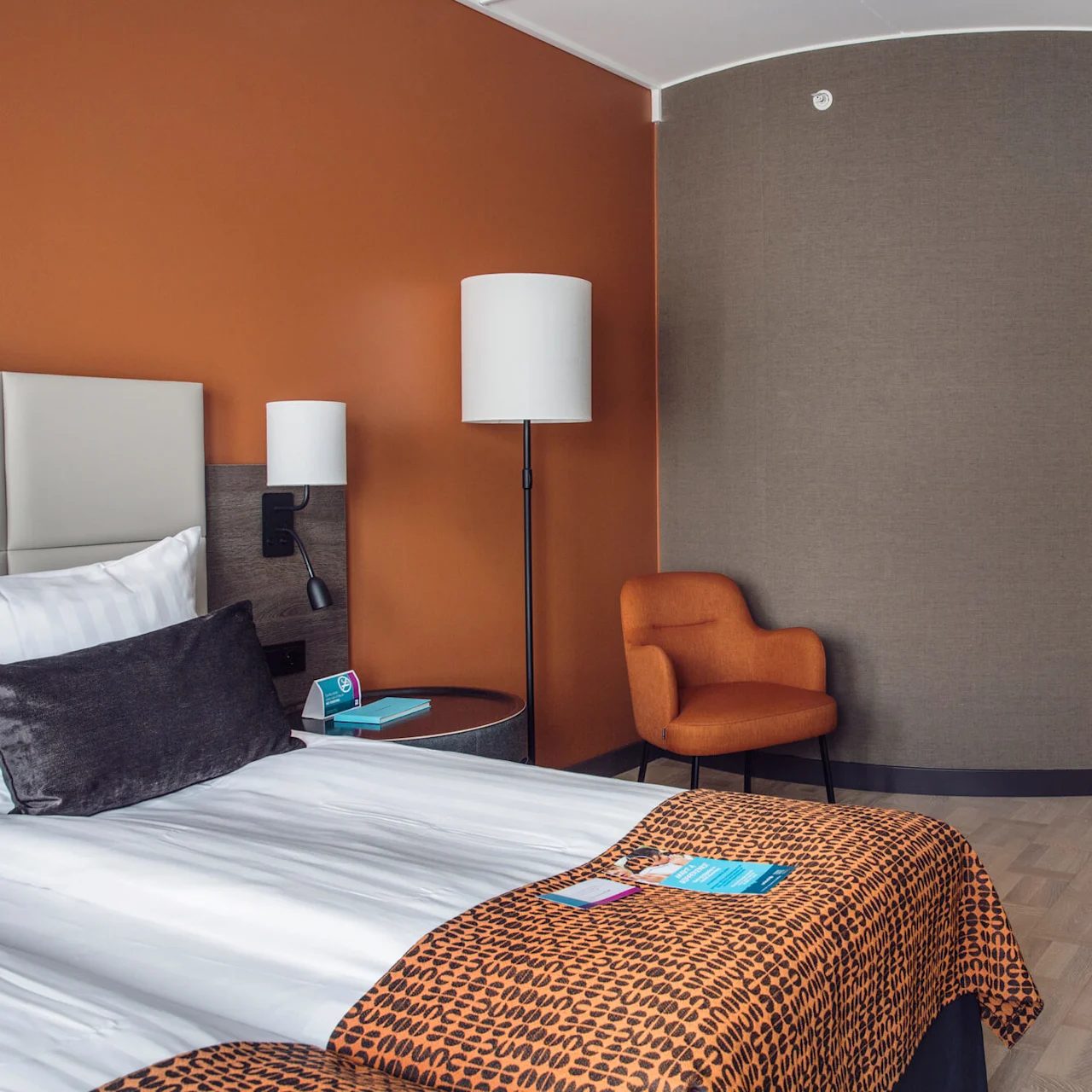 Säng med vita lakan och orangefärgad mönstrad filt vilar bredvid ett runt bord och lampa, i ett modernt hotellrum med en orange stol och trägolv.