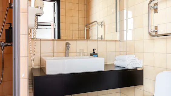 Ett modernt badrum med en diskho med en elegant kran på en svart bänk, beige kaklade väggar, en hårtork och prydligt vikta handdukar bredvid handfatet.