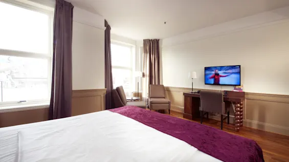 Et godt belyst hotellrom med en stor seng, TV på, stol ved vinduet, og tregulv.