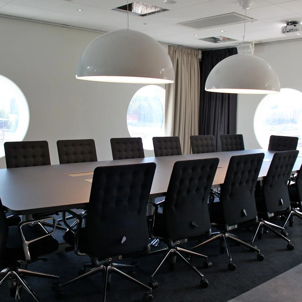Et langt konferencebord med sorte stole omgiver et tomt rum. To store kuppellamper hænger over, og cirkulære vinduer giver udsigt til udenfor.