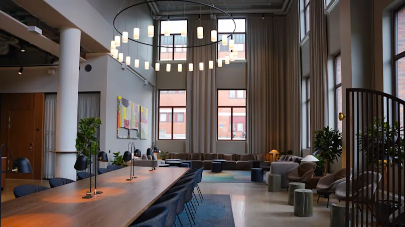 A modern office lounge area with sofas, a long wooden table with chairs, and hanging lights against large windows.