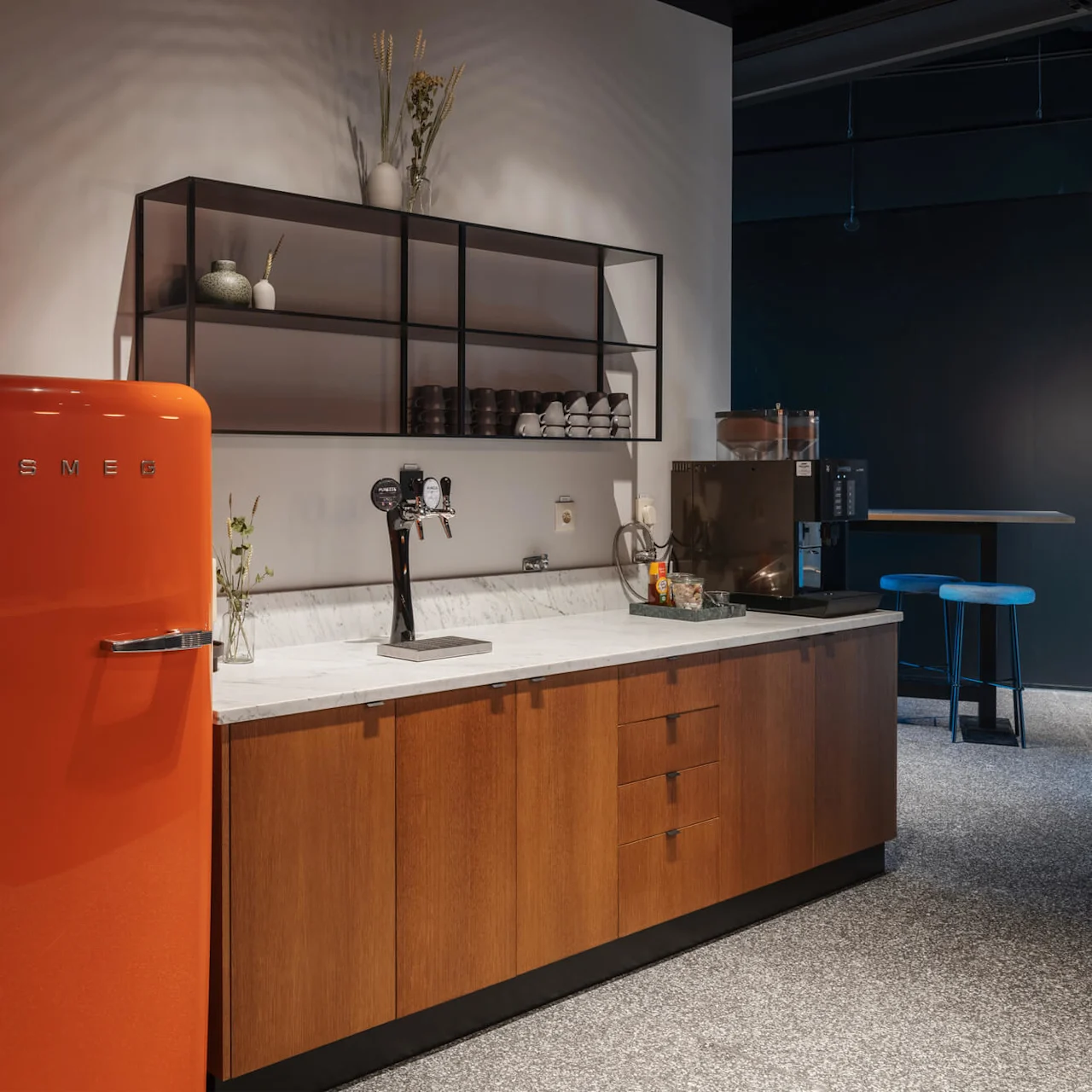 A retro orange SMEG refrigerator stands beside a modern kitchen counter with a marble top, shelves with dishes, a coffee dispenser, and a tray of snacks in a stylish interior.