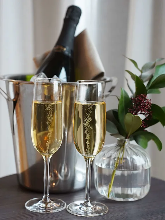 Champagne i glass med en flaske i en isbøtte og en vase med grønt, plassert på et trebord mot draperte gardiner.