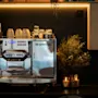 En koselig kaffestasjon med en maskin, kopper, kaffekapsler, tørkede blomster og tente lys. Et BRANNSENT-skilt er synlig.