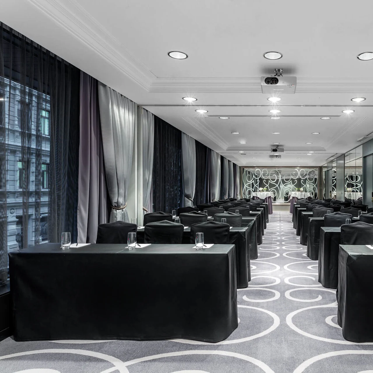 Tables are set with water glasses and notepads, arranged classroom-style in a conference room with patterned carpet, large windows with curtains, and mirrored walls enhancing the elegant ambiance.