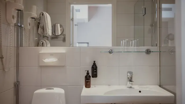 Handfat med kran i ett vitkaklat badrum; flankerad av tvåltvålpumpar och glas. I närheten finns en toalett, väggmonterad hårtork och handdukshängare som förstärker hotellets enkla, funktionella estetik.