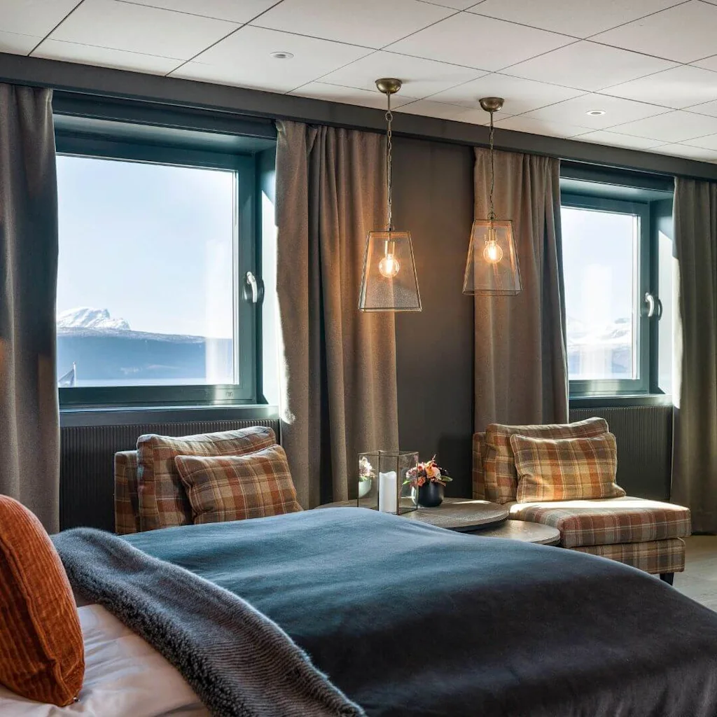 A cozy hotel room features a large bed with burnt orange pillows, facing two windows with mountain views. Two pendant lights hang above plaid chairs, with a wall-mounted TV nearby.