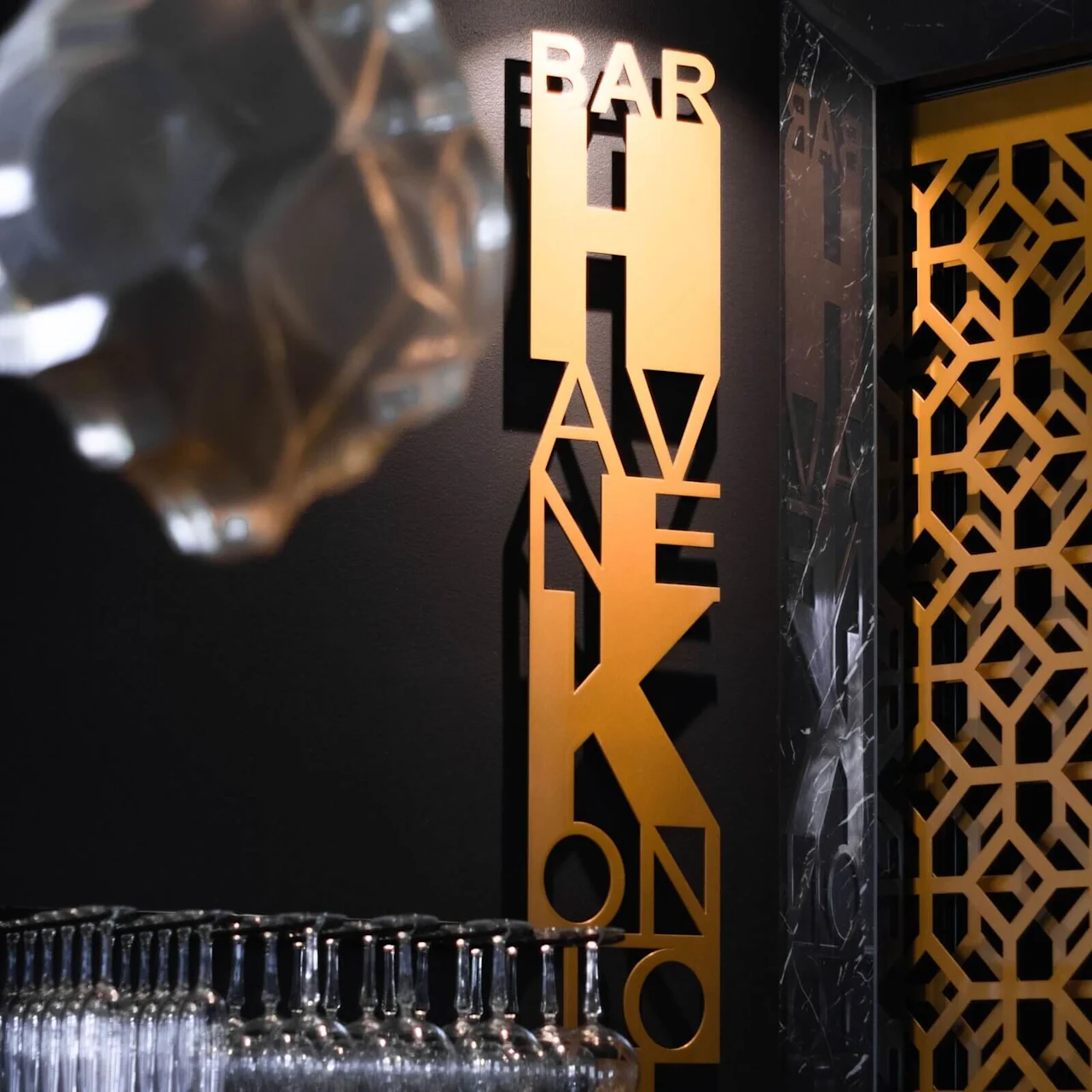Golden signage reading "BAR HAVNEKONTOR" beside an array of upside-down glasses on a shelf, suggesting a bar setting.