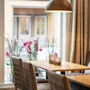 Et træbord omgivet af polstrede stole, med to stearinlys, står under en hængende lampe. Lyserøde blomster i vaser står ved vinduet, med udsigt over en gade.