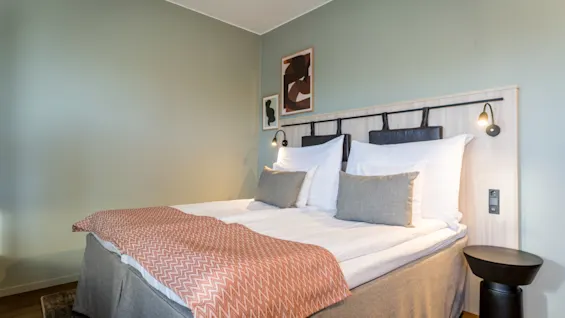 Bed with white sheets and pillows, adorned with a pinkish patterned throw, stands against a light green wall featuring abstract art, flanked by modern side tables and lamps.