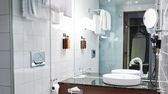 A modern bathroom with a toilet and sink. Towels hang above a counter with a round mirror. Glass shelves hold toiletries. Walls and floor are tiled.