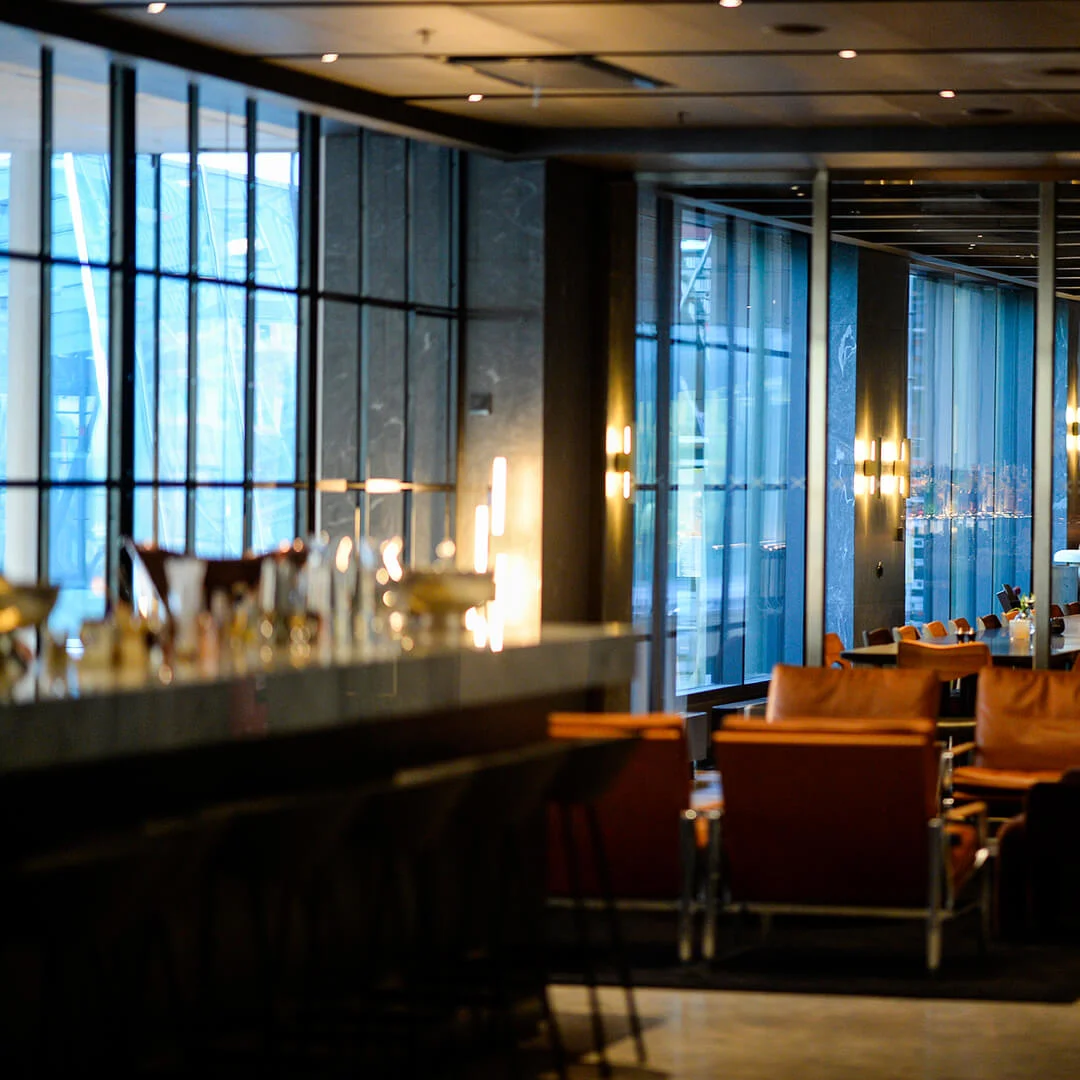 Stoler og bord er arrangert i en svakt opplyst, moderne restaurant med store vinduer og elegant innredning, som skaper en sofistikert spisestemning.