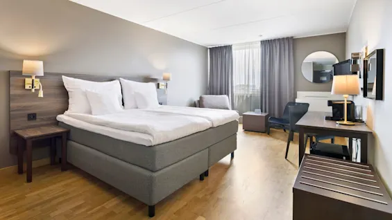 Et pænt og velordnet hotelværelse har en dobbeltseng med hvidt linned, et træbord med en lampe og et fladskærms-TV. Gardiner dækker et vindue, med neutrale farver på vægge og gulv. 