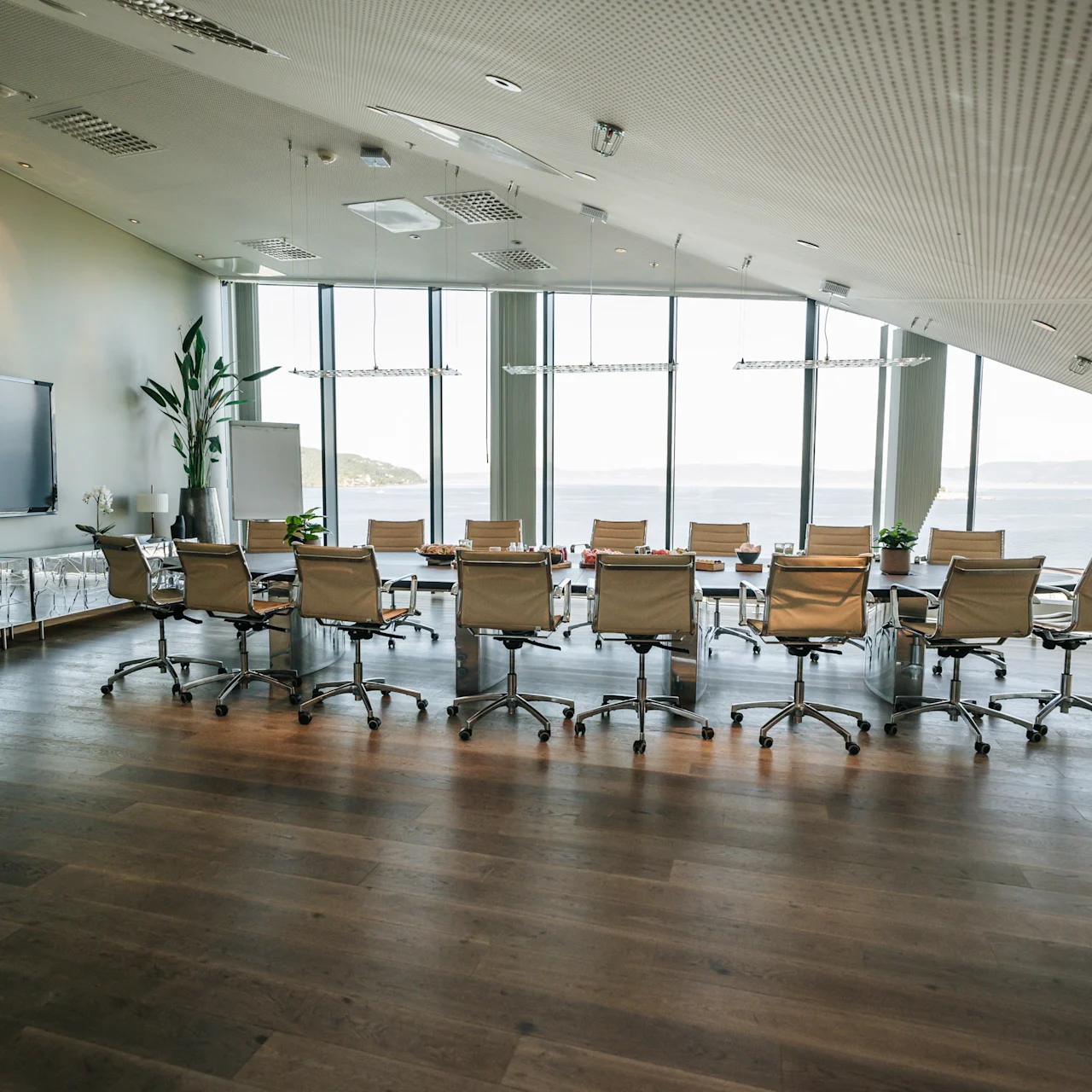 A large conference table with swivel chairs sits in a modern meeting room. Floor-to-ceiling windows offer a waterfront view. The room has plants, a TV, and a curved ceiling.