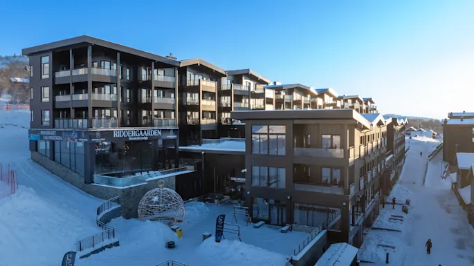 Riddergaarden Mountain Lodge facade in winter, with snow-covered grounds. Visible text: Casa Montagna, Casa Moritz. People walking.