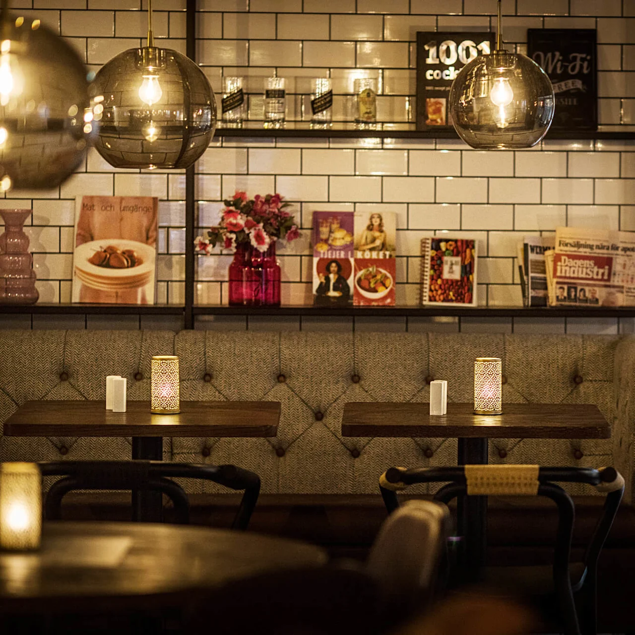 A cozy café interior with upholstered bench seating, hanging lights, and decorative wall tiles displaying posters and newspapers.