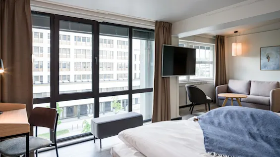 A neatly made bed with a blue throw sits in a modern hotel room. A TV is mounted, alongside a desk and chair. Large windows reveal an urban scene outside.