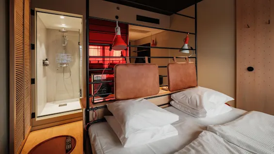 A modern hotel room features a neatly made bed with white pillows and a shower visible through a glass wall. Two lamps hang overhead in a stylish, minimalistic design.