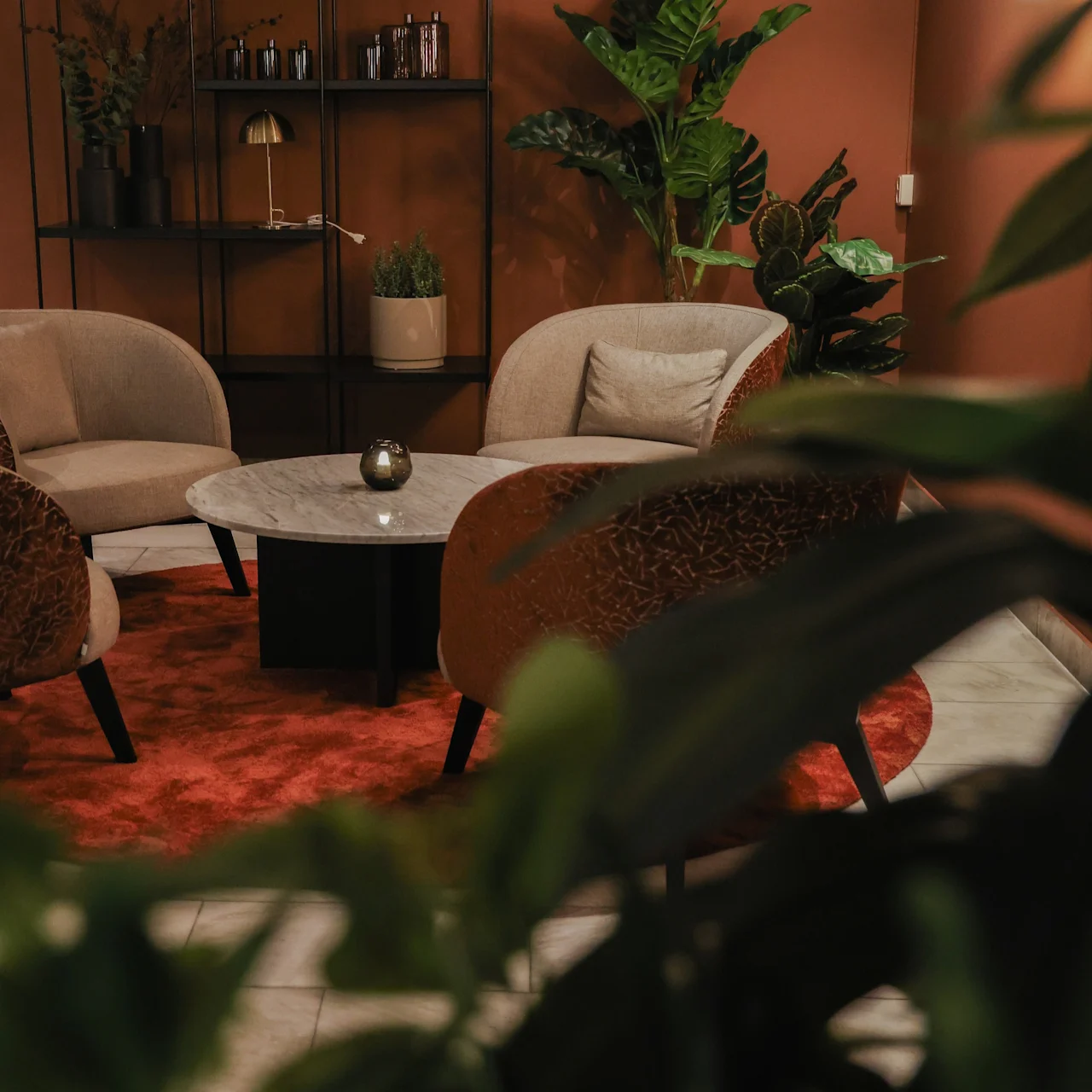 A round marble table is surrounded by beige chairs on a red rug, in a cozy room with plants and a brown wall-mounted shelf.