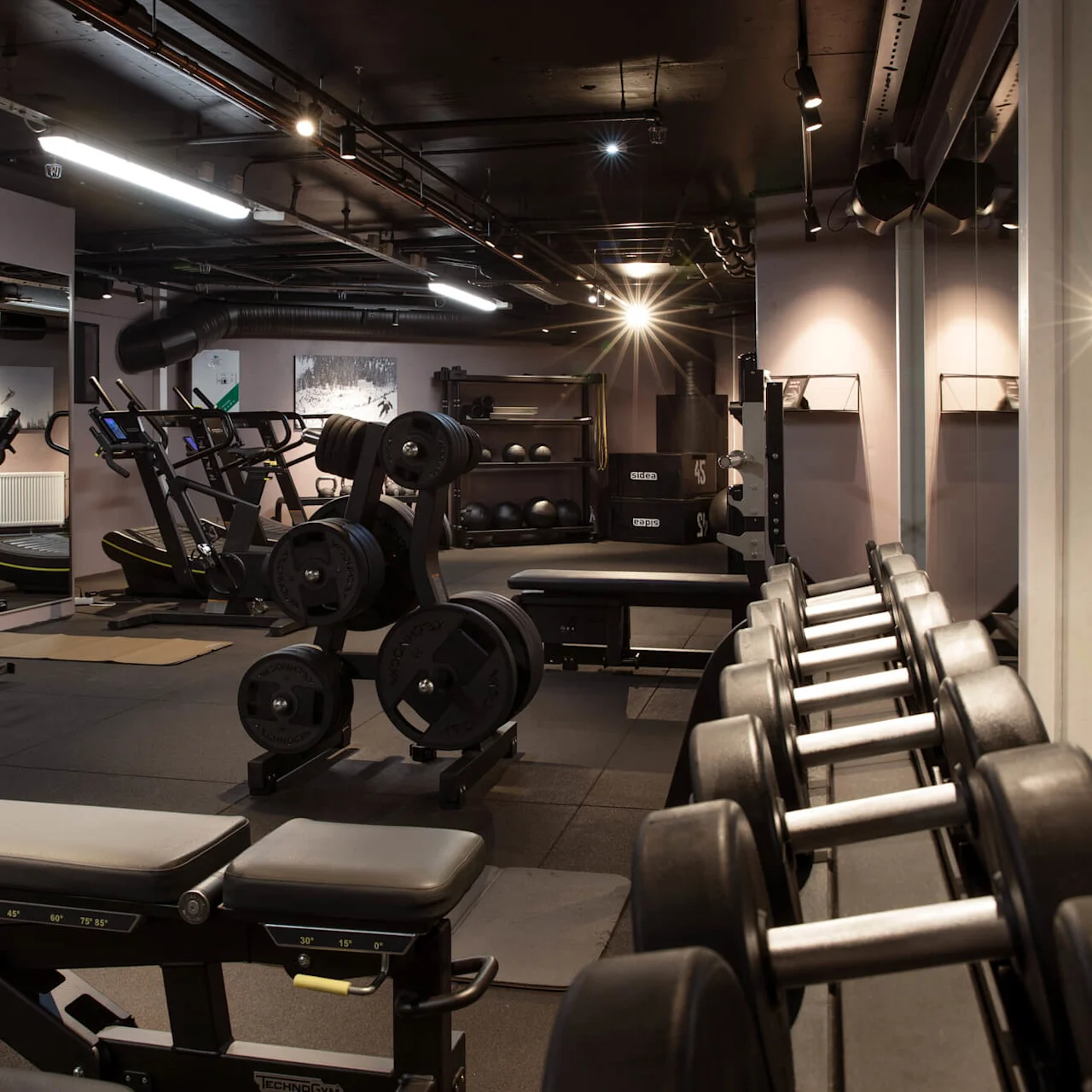 A gym with benches and dumbbells in the foreground, exercise bikes and treadmills in the background, under bright ceiling lights and surrounded by mirrors.