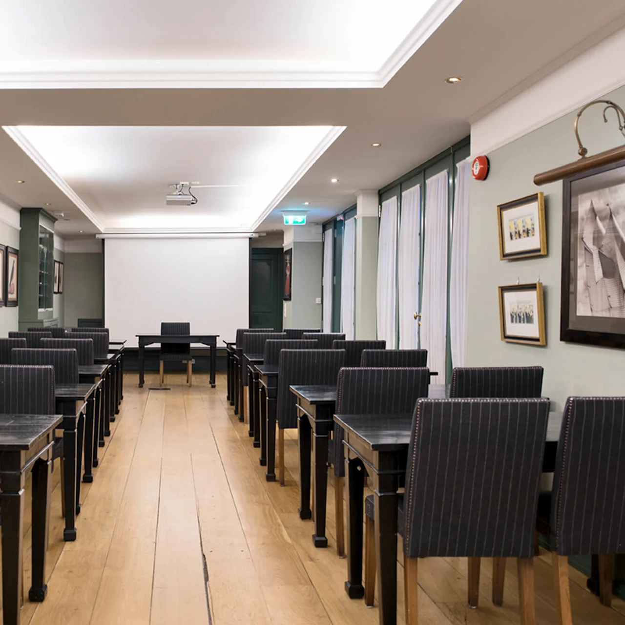 Rekker av svarte stoler vendt mot et lerret i et tomt konferanserom med innrammede bilder på veggene.