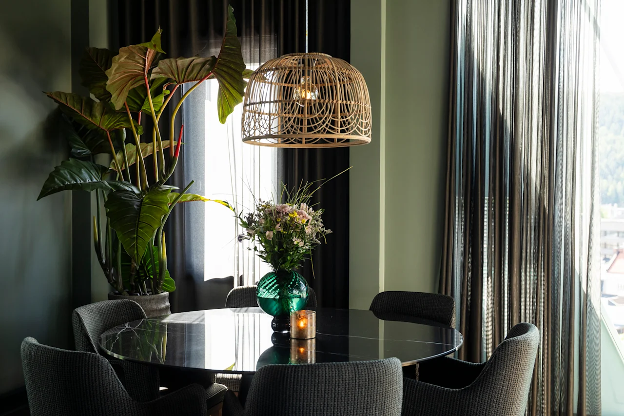 Et stilig spiseområde med et rundt svart bord, fire stoler, en stor plante, en taklampe i rotting, blomster og et lys.