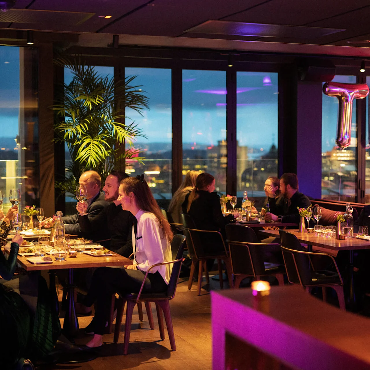 People dine and socialize at elegantly set tables in a stylish restaurant overlooking a cityscape at dusk. Shiny balloon letters spell "THE" against large windows, with ambient lighting.