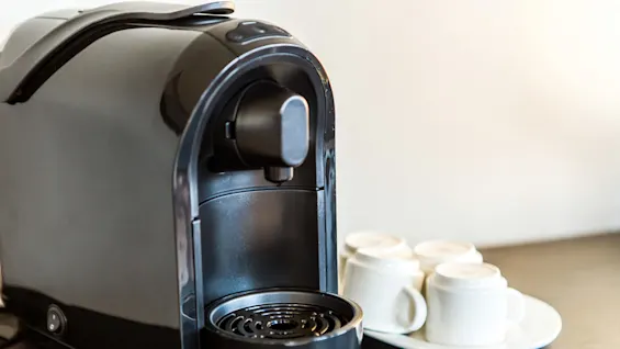 A sleek, black coffee machine sits on a dark countertop beside a white platter holding four upside-down ceramic cups, indicating preparation for brewing coffee.