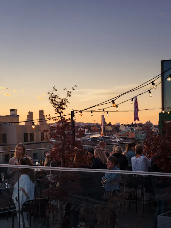 Gjester nyter drinker og samtaler på en livlig takbar ved solnedgang, med utsikt over byen. TAK på Hobo i Stockholm.
