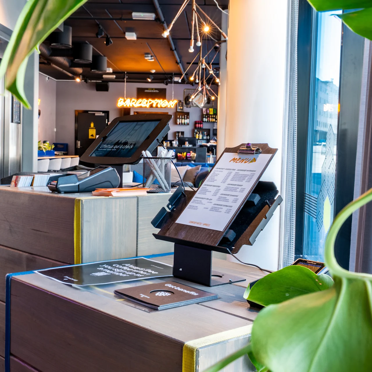 Digitaalinen sisäänkirjautumisautomaatti, jossa on näyttö ja leikekansio, sijaitsee puupöydällä modernissa hotellin aulassa. Lähellä loistaa neonkyltti "RECEPTION" vastaanottotiskin yläpuolella.