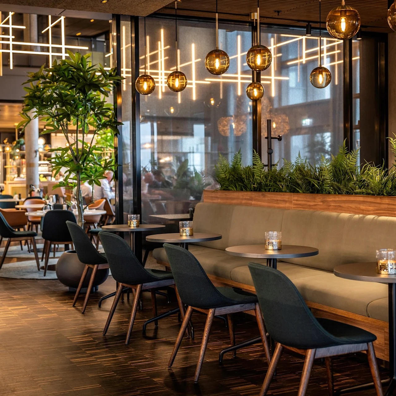 Elegant siddeområde med grønne polstrede stole og træborde; stemningsfuld belysning og indendørs planter skaber en hyggelig, stilfuld atmosfære i en moderne restaurantindstilling.
