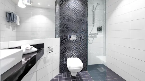 Et moderne badeværelse med et flydende hvidt toilet mod en mosaikflisevæg. En glasindkapslet bruser med krombeslag står ved siden af, omgivet af hvide fliser.