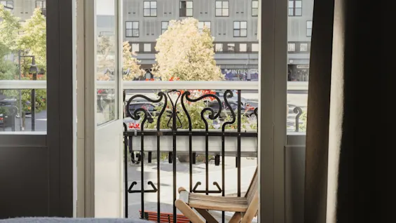 Utsikt från en hotellrumsbalkong med en trästol, med utsikt över en gata med träd och byggnader. Synlig text: INTERSPORT.