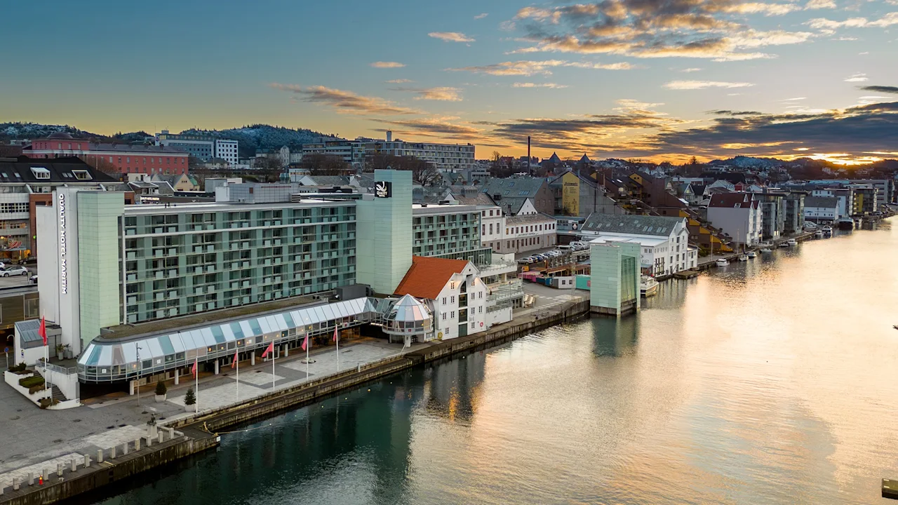 A large waterfront hotel stands near a calm canal, surrounded by a row of colorful buildings. The setting sun casts a golden glow over the scenic urban landscape.