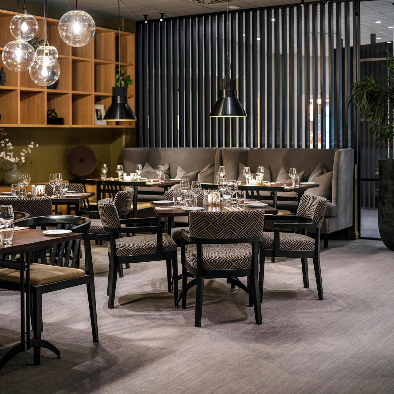 Tables set for dining with patterned chairs, surrounded by wooden shelving and potted plants; pendant lights illuminate the modern restaurant interior, creating a warm and inviting atmosphere.