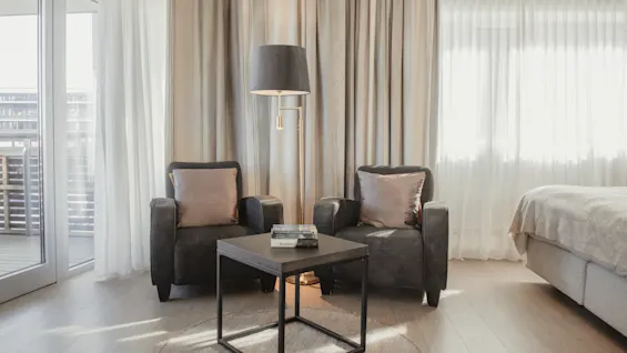 Ett mysigt hotellrum med två fåtöljer, ett litet bord med böcker, en golvlampa och en säng, badat i naturligt ljus.