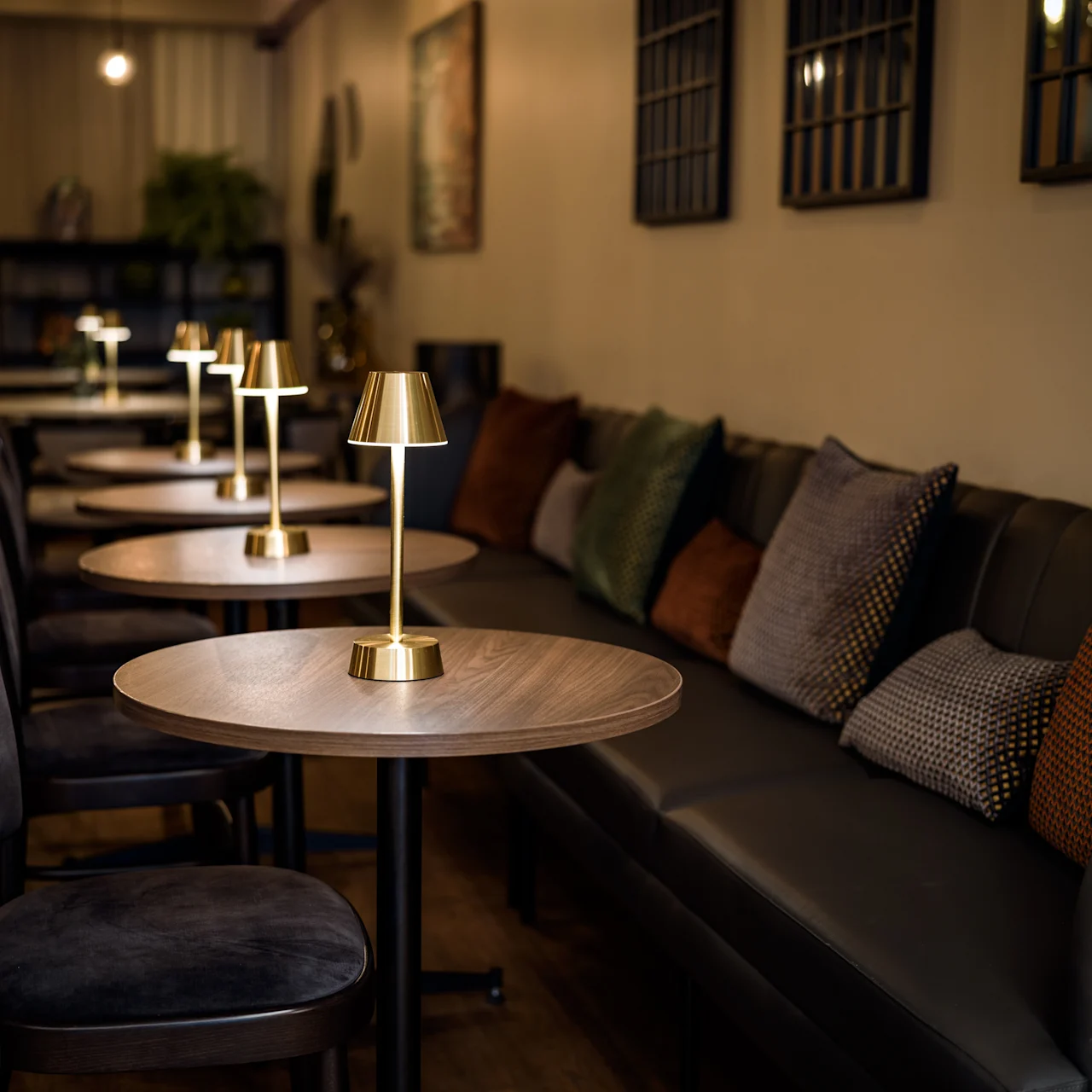 Golden table lamps illuminate round wooden tables in a cozy, dimly-lit café with cushioned seating. Soft pillows line the benches against the warm-colored walls, creating an inviting atmosphere.
