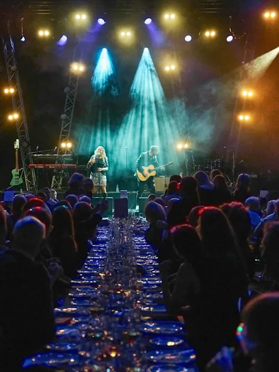 Et liveband opptrer på en lyst opplyst scene for et publikum som sitter ved bord, noe som skaper en energisk atmosfære.