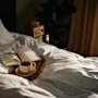 En bog og briller hviler på en sengbakke med en rygende kaffekop, omgivet af hvidt sengetøj i et hyggeligt soveværelse, blødt oplyst af naturligt lys.