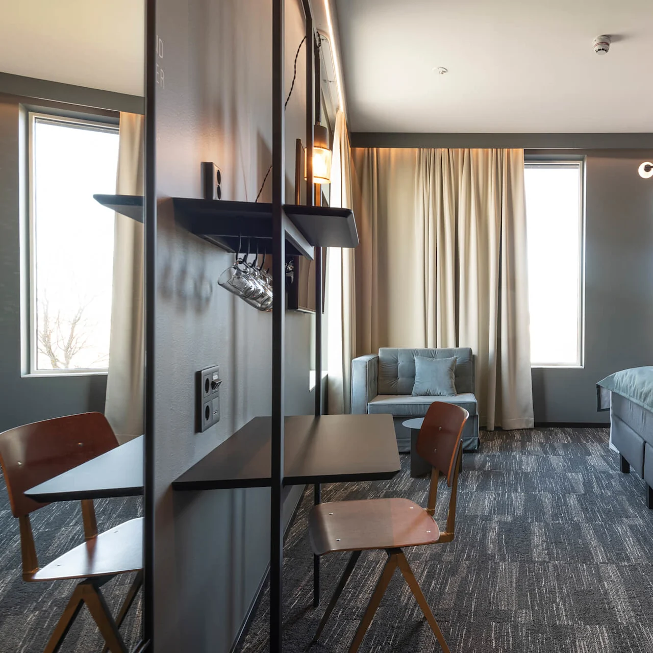 A modern hotel room features a bed with a green blanket, a small desk, and a chair. Soft lighting and large windows create a welcoming atmosphere. Text: "GYM & FRONTDESK EN 24/7 DON'T FORGET TO."