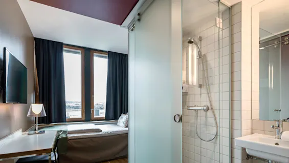 A modern hotel room with a bed by large windows, a desk with a lamp and chair, and an adjacent bathroom with a shower and sink separated by frosted glass.
