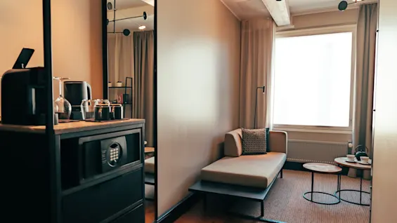 Ett mysigt hotellrum med en schäslong vid ett ljust fönster och en minibar med kaffemaskin och kassaskåp.