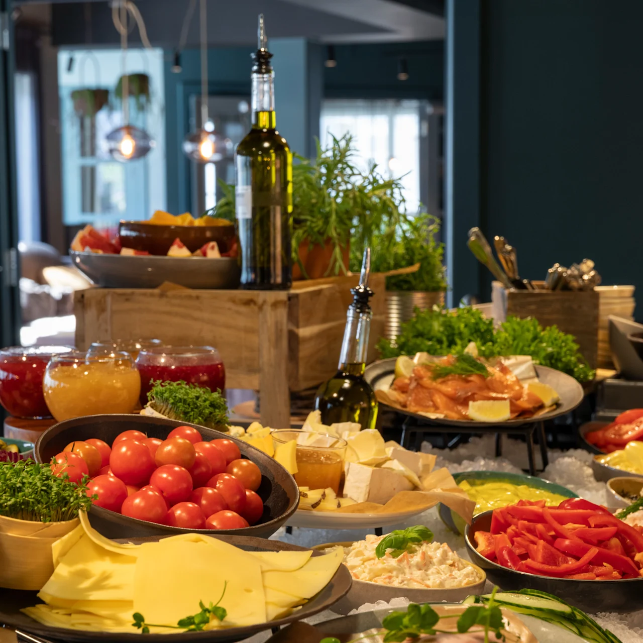 Et udvalg af friske grøntsager og oste på et buffetbord. Omgivet af urter, oliefalder og forskellige retter, opstillet i en hyggelig spisestue.